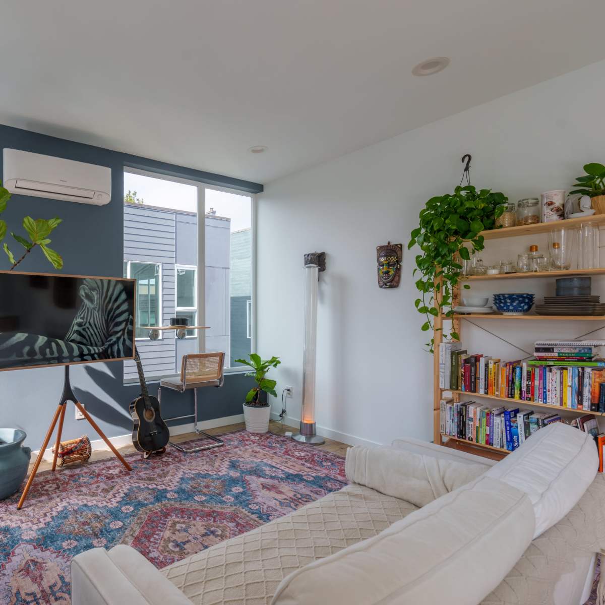A bright living room featuring a large television, a white couch, a bookshelf filled with books, and several indoor plants.