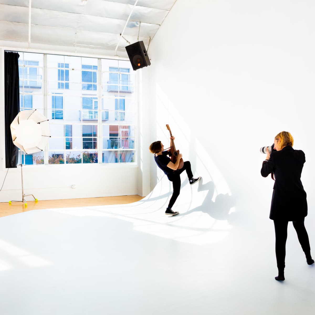 A person plays guitar against a white wall while another person photographs them in a brightly lit studio.