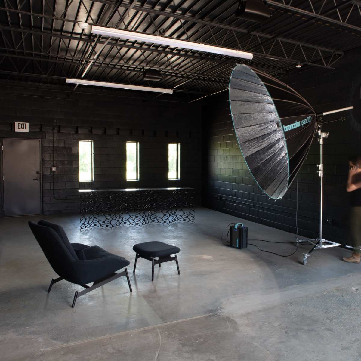 A dimly lit studio with black walls featuring a large umbrella light setup, a compact chair, and a small table.