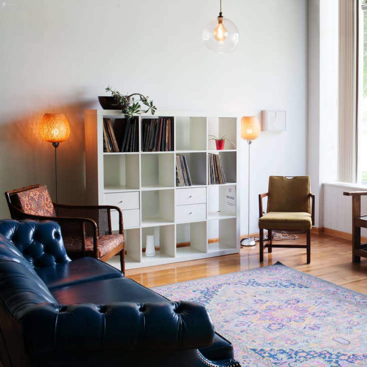 A well-lit living room features a blue leather sofa, a wooden bench, a shelving unit filled with records, and a large window revealing greenery outside.
