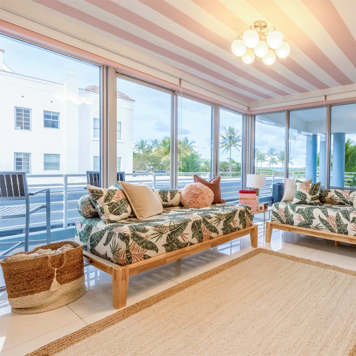 A bright living space features two beds with tropical-patterned bedding, large windows overlooking palm trees, and a light-colored area rug on a glossy floor.