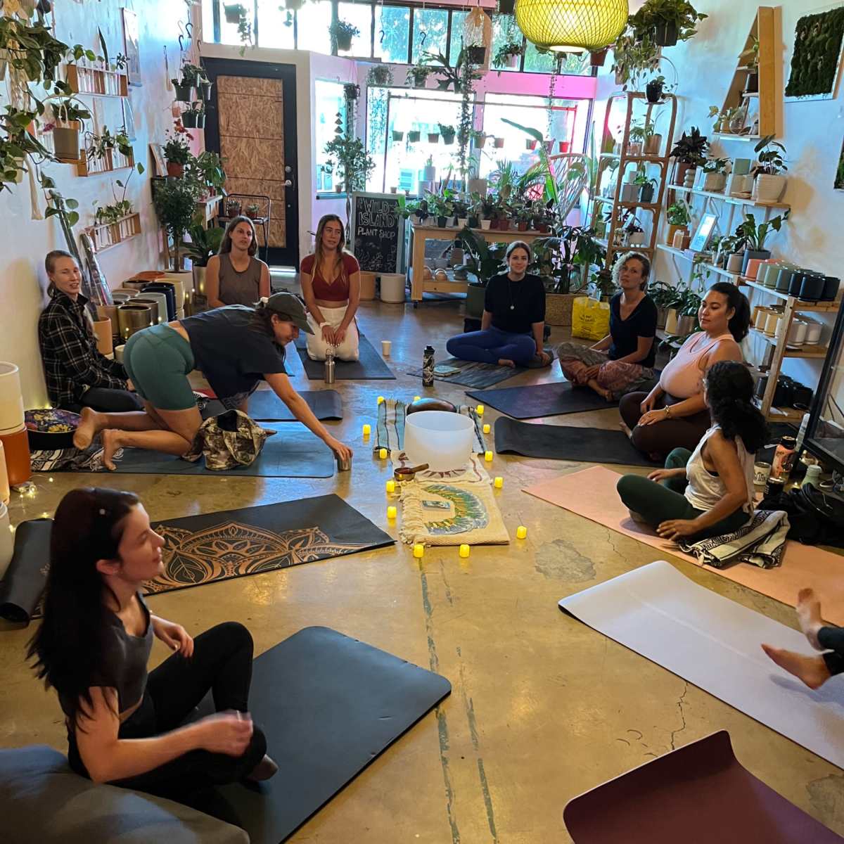 A group of individuals participates in a yoga or meditation class in a plant-filled studio, seated on mats arranged around a central layout of candles and a bowl.