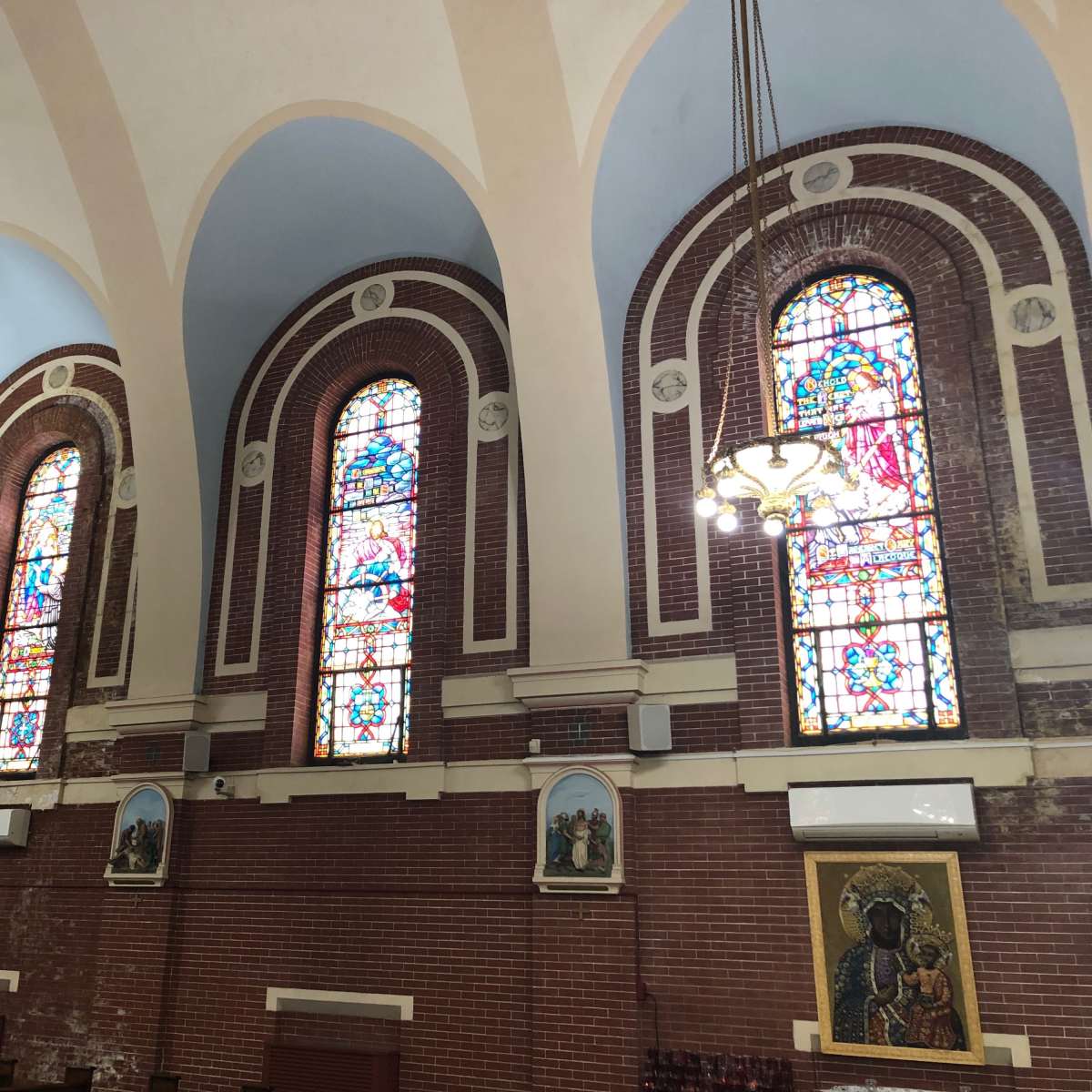 The interior of a church, featuring arched stained glass windows and decorative lighting fixtures along the walls.