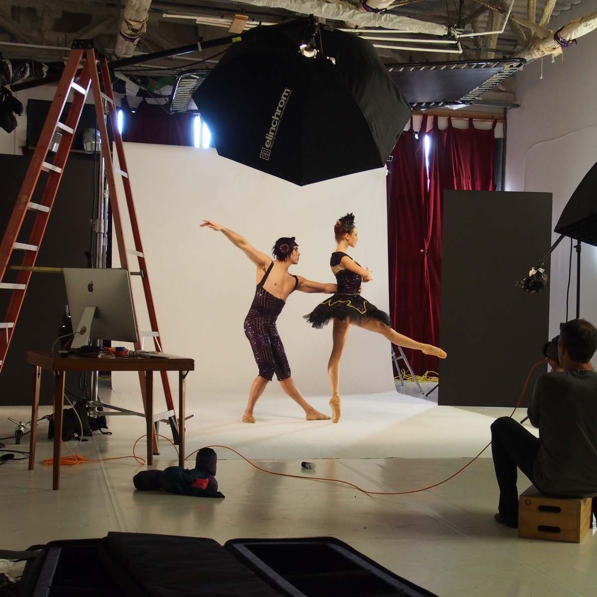 Two dancers are posed on stage in a photography studio with lights and equipment set up, while a photographer captures their performance.