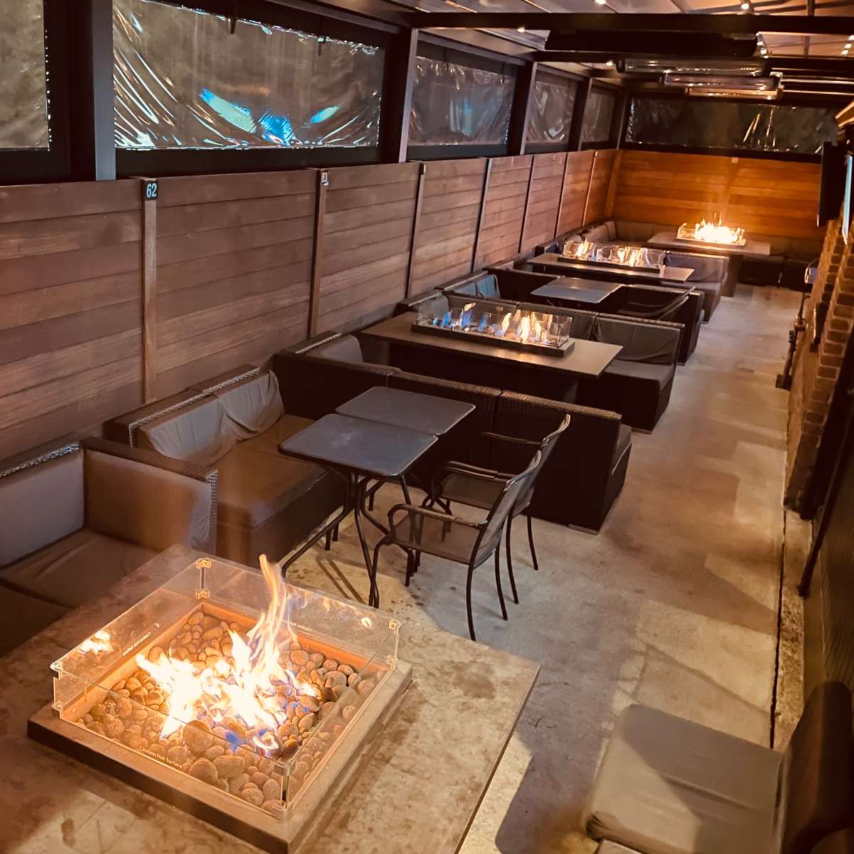 A modern outdoor lounge with several tables featuring fire pits, surrounded by wooden walls and a ceiling.
