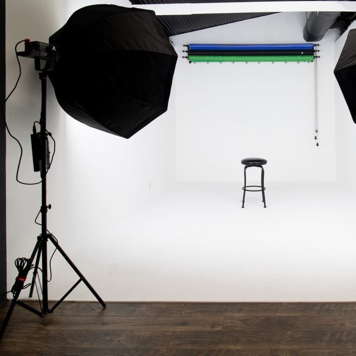 A photography studio features a white backdrop, a black stool in the center, and large softbox lights positioned on either side.