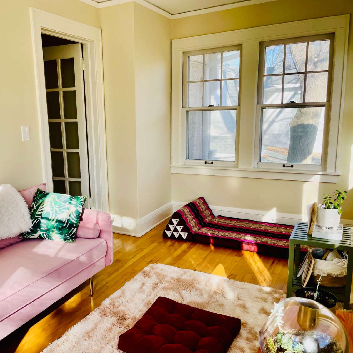 A bright living room features a pink sofa with decorative pillows, a light-colored rug, and large windows allowing sunlight to stream in.