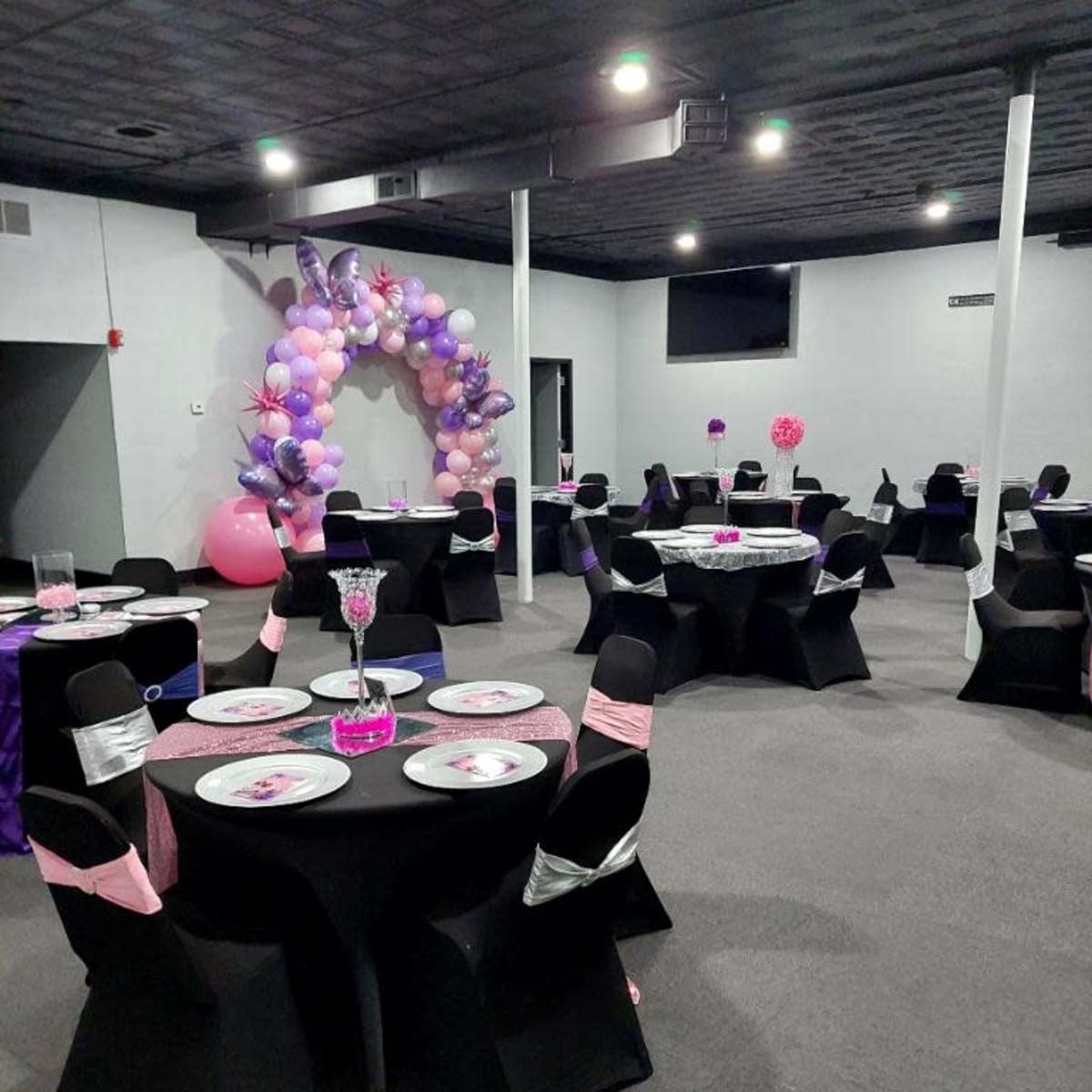A party venue with black tables adorned with pink and purple decorations, surrounded by balloon arches and centerpieces.
