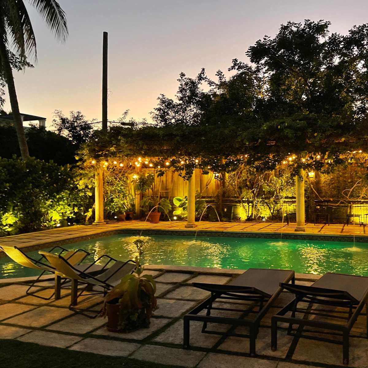 A pool area surrounded by lounge chairs and illuminated by string lights, set against a twilight sky.