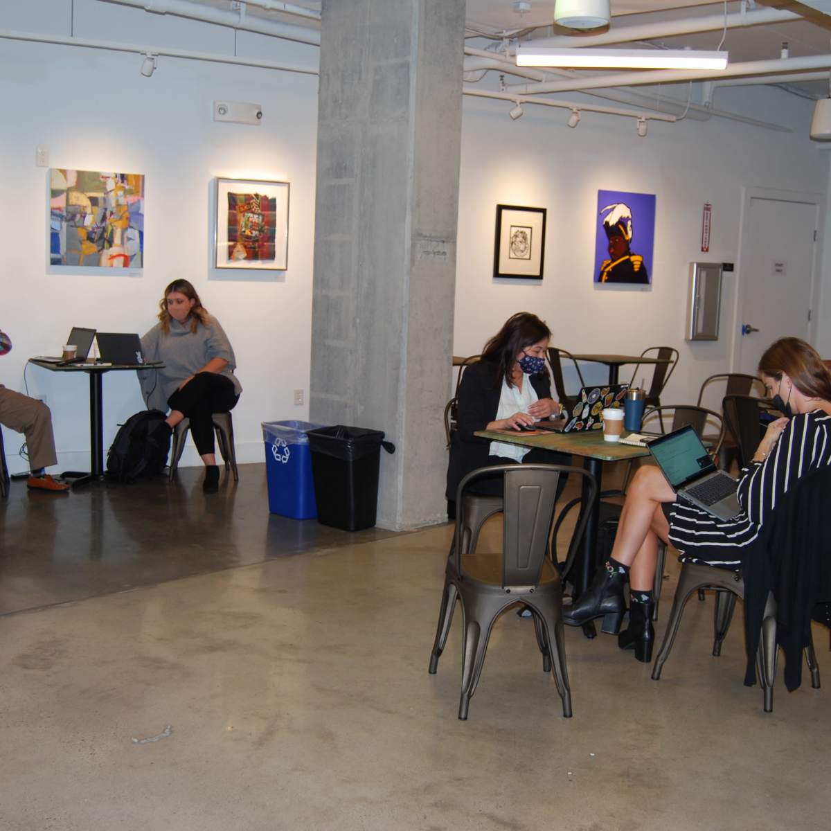 A spacious room with several people working at tables, wearing masks, and various artworks displayed on the walls.