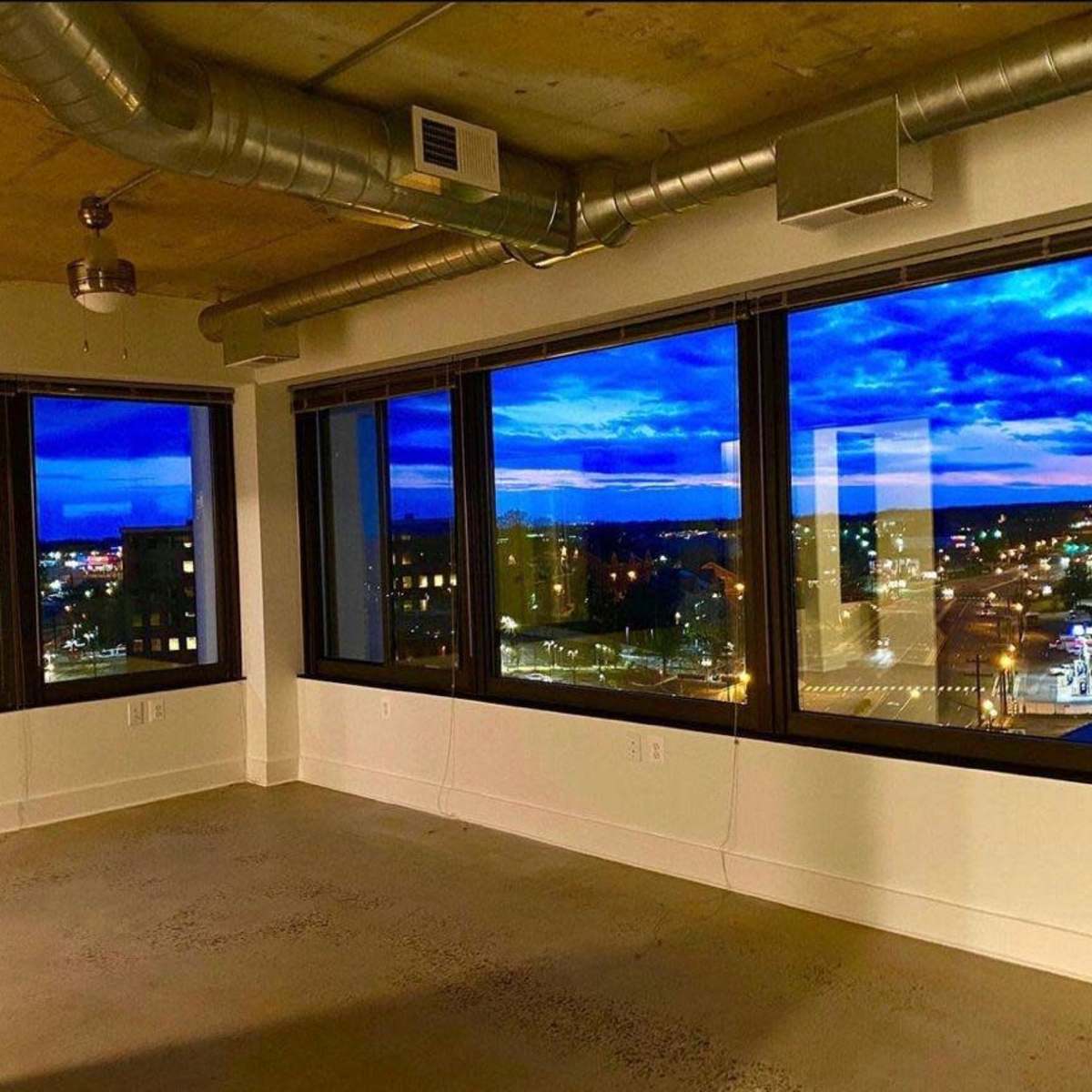 A vacant room with large windows overlooking a cityscape illuminated by evening lights, against a backdrop of a blue sky filled with clouds.