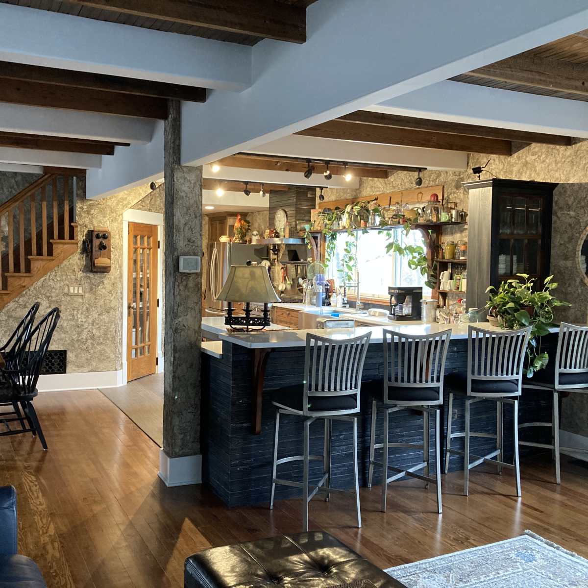 A spacious kitchen and dining area features wooden beams, a central island with bar stools, and a staircase leading to an upper level.