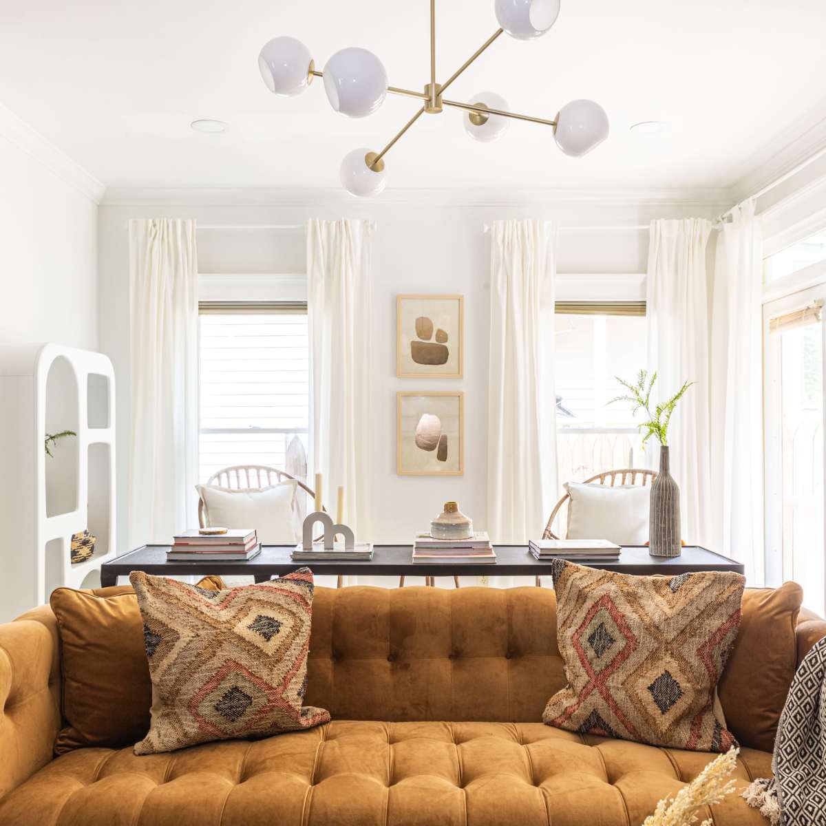 A living room with a brown tufted sofa, patterned cushions, a modern chandelier, and large windows dressed with sheer curtains.
