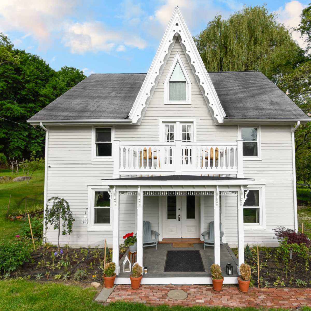 A white, three-story house with a peaked roof, a front porch, and a grassy yard surrounded by trees.