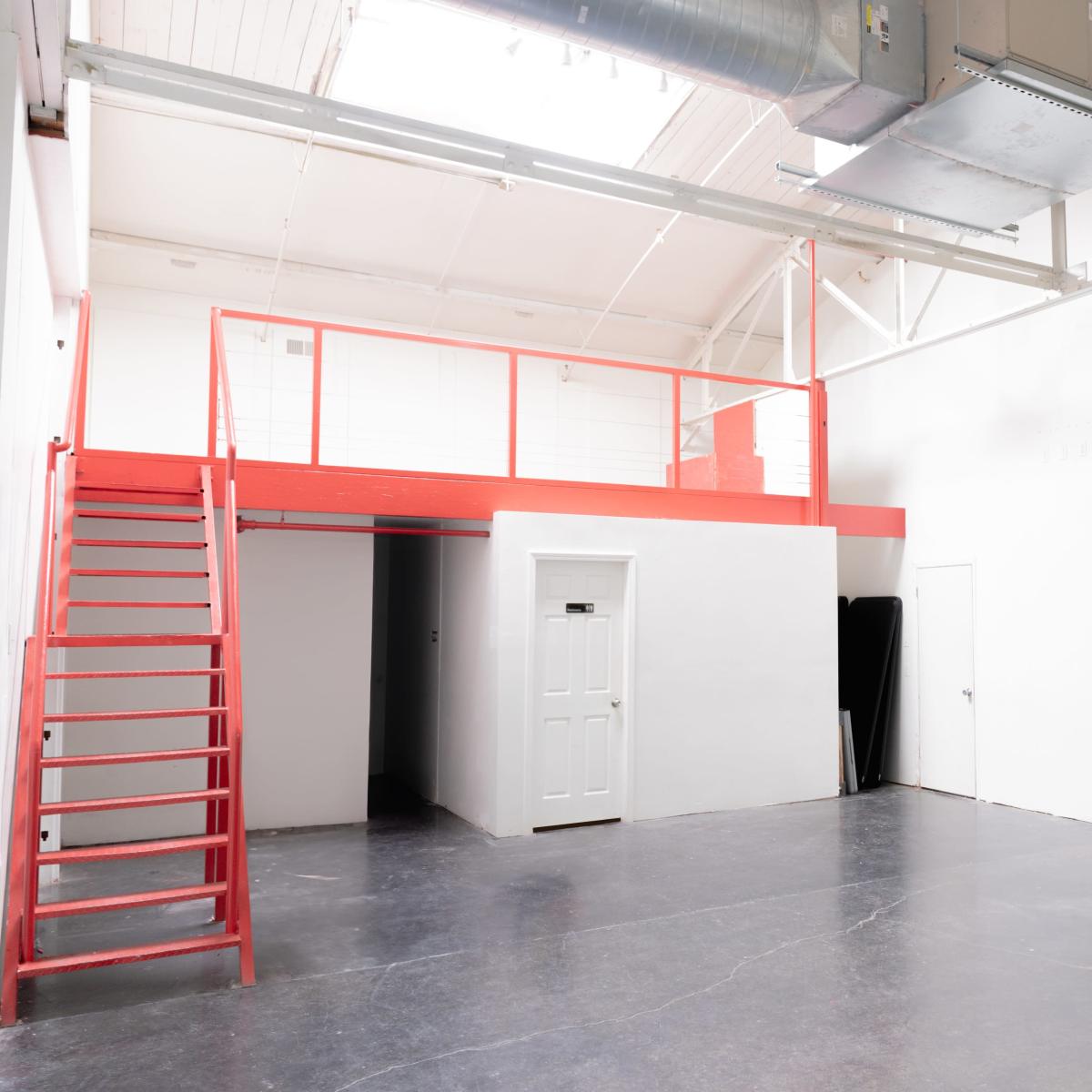 An industrial-style interior space featuring a red staircase on the left leading to a loft area, a yellow spiral staircase on the right, and white walls with polished concrete flooring.