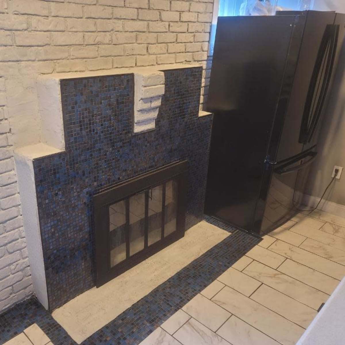A white brick wall with a tiled fireplace and a black refrigerator beside it in a kitchen setting.