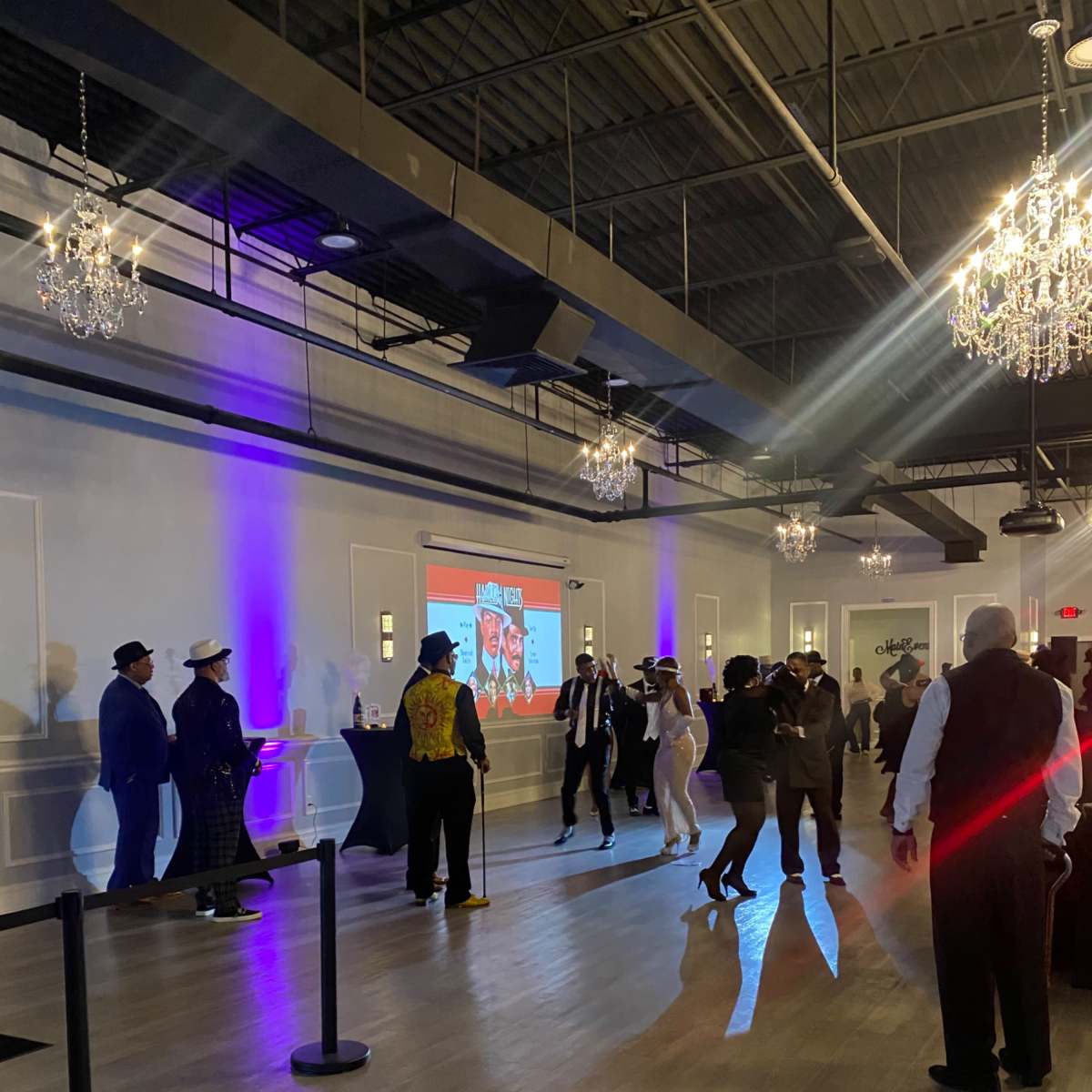 A group of people dressed in formal attire gathers in a spacious ballroom with chandeliers, while a screen displays images in the background.