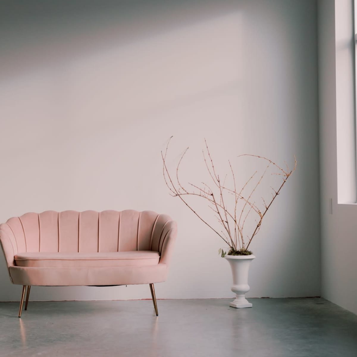 A pink scalloped sofa is placed against a light wall, accompanied by a slender vase with bare branches and two gold candle holders on the floor.