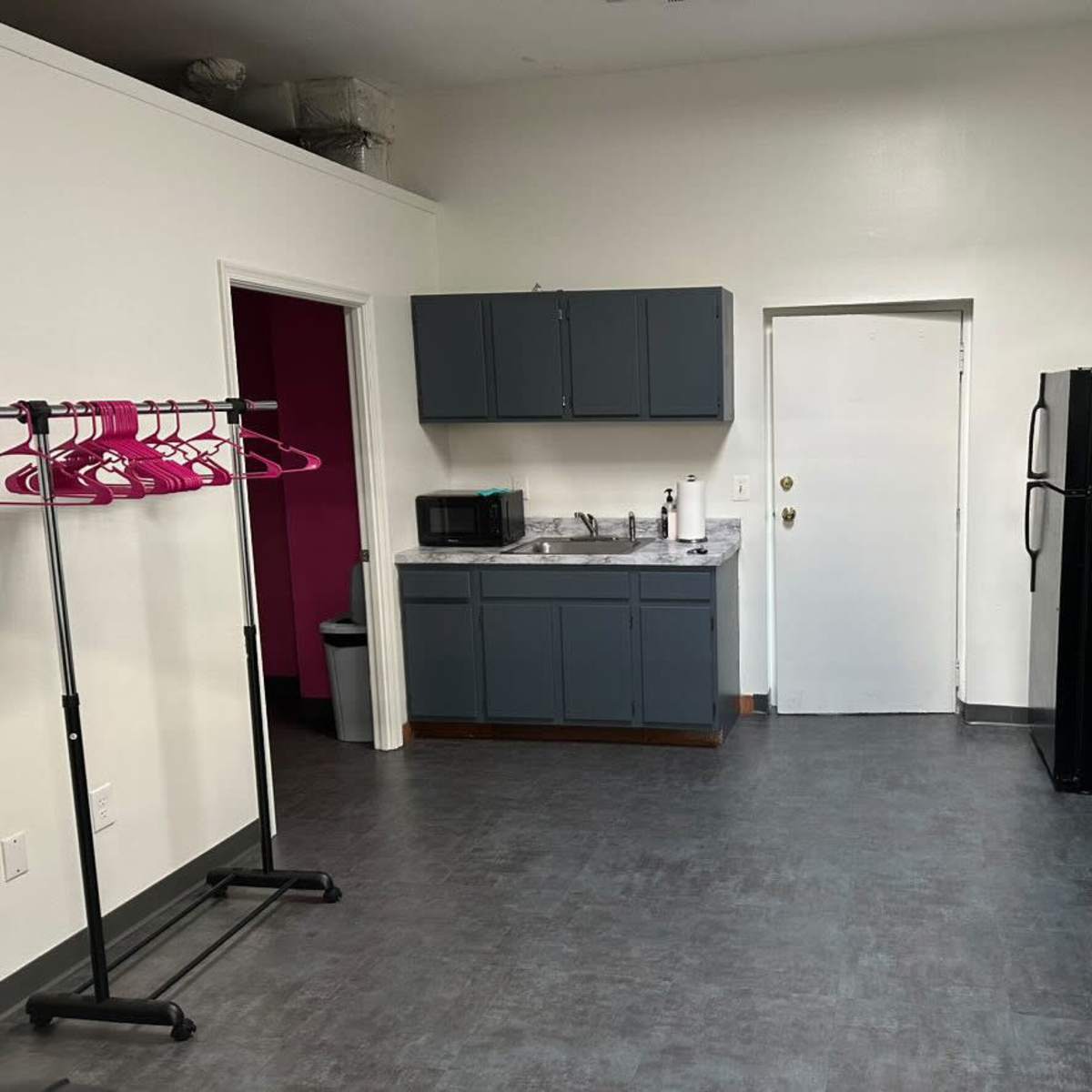 A sparse room with gray flooring, a kitchenette along one wall, a black refrigerator, a clothing rack with pink hangers, and a door leading to a bathroom.