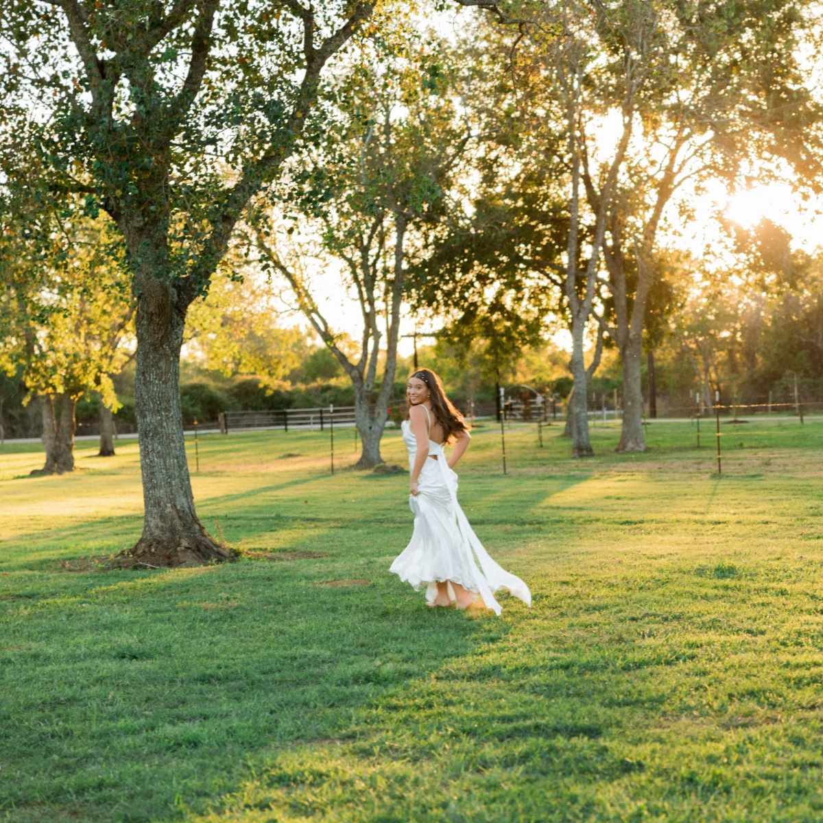 A woman in a white dress runs across a grassy field at sunset, surrounded by trees and a fenced area.