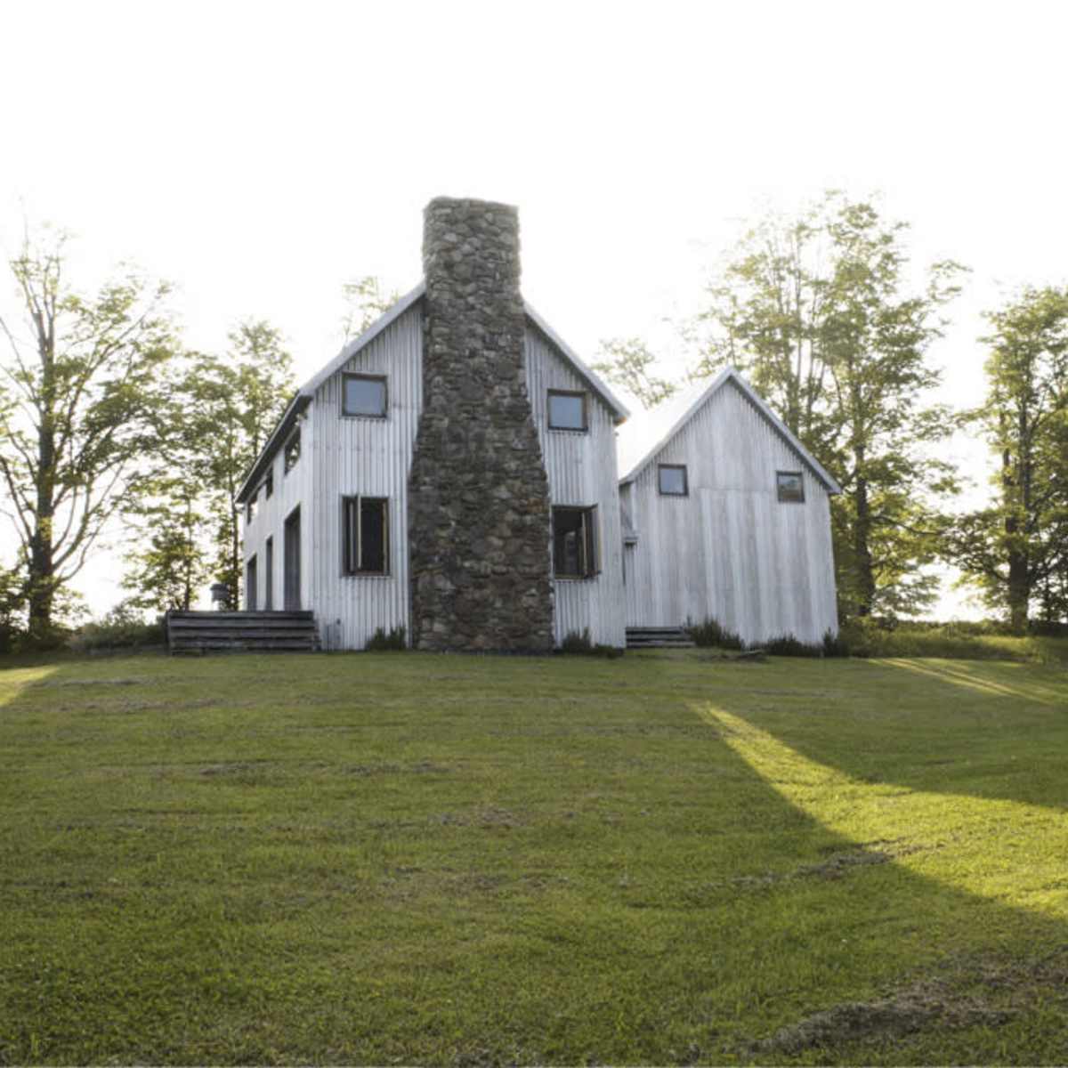A two-story, white house with a stone chimney stands amidst a grassy area and trees.