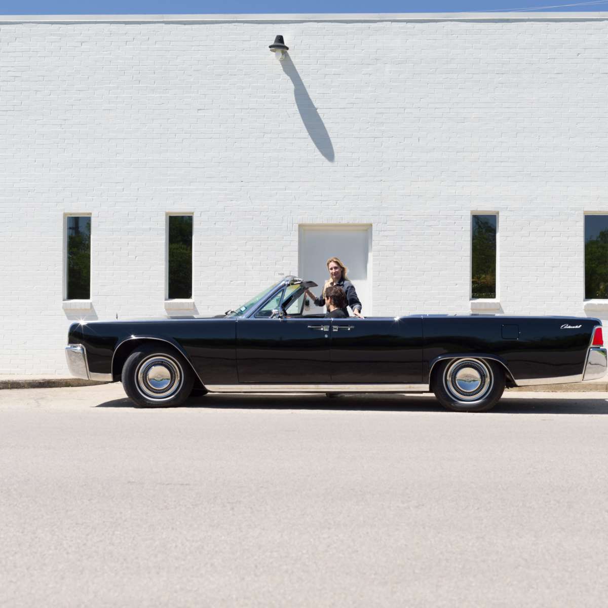 A black convertible car is parked in front of a white building with small rectangular windows, while a person stands beside it.