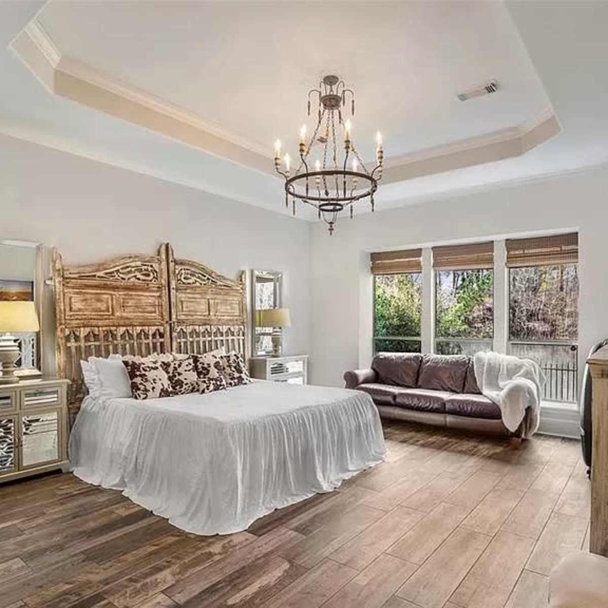 A spacious bedroom featuring a wooden bed frame, large windows with blinds, and a mix of light and dark furniture.