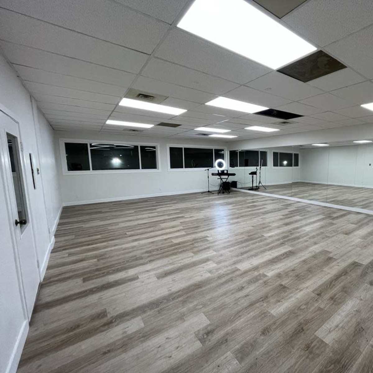 A spacious, empty room with wood-like flooring, white walls, a large mirror on one side, and several ceiling tiles missing.