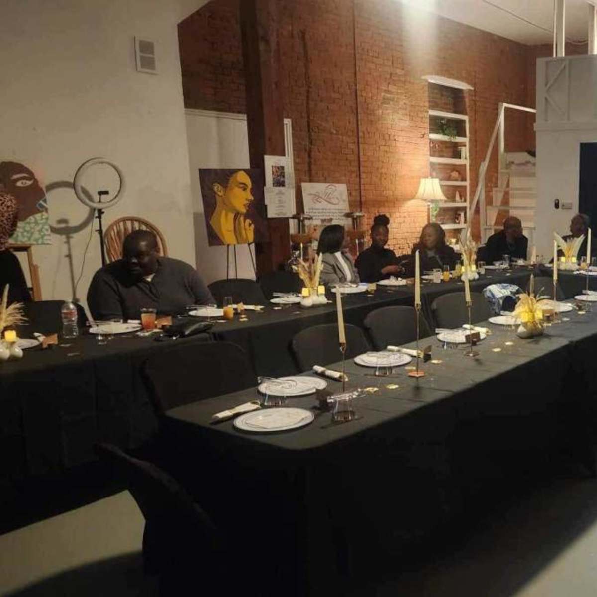 A long dining table is set with plates and candles in an art-filled space, with several people seated along one side and a person standing in the background.