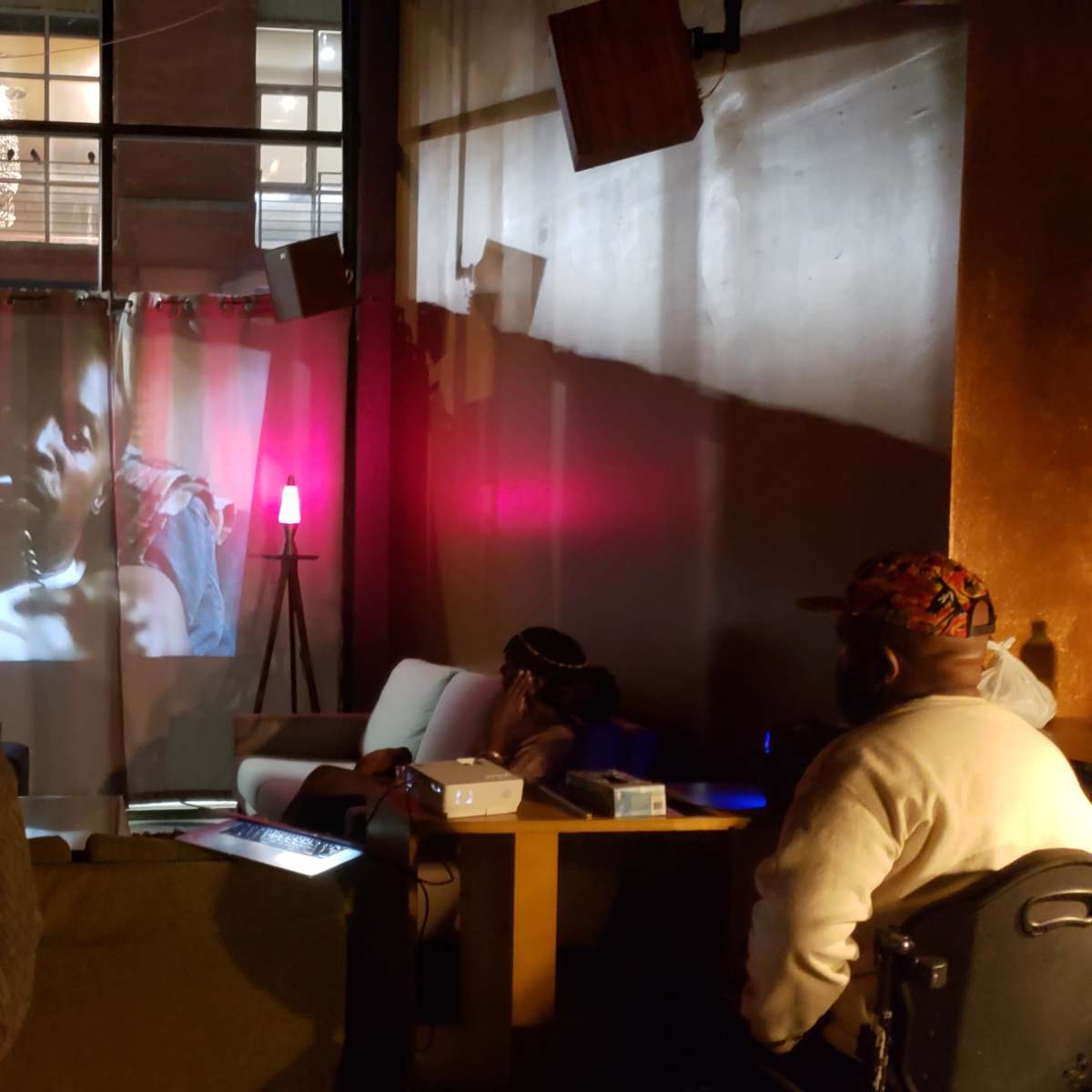 A person stands in a dimly lit room watching a film projected onto a curtain, while another person sits on a couch nearby.