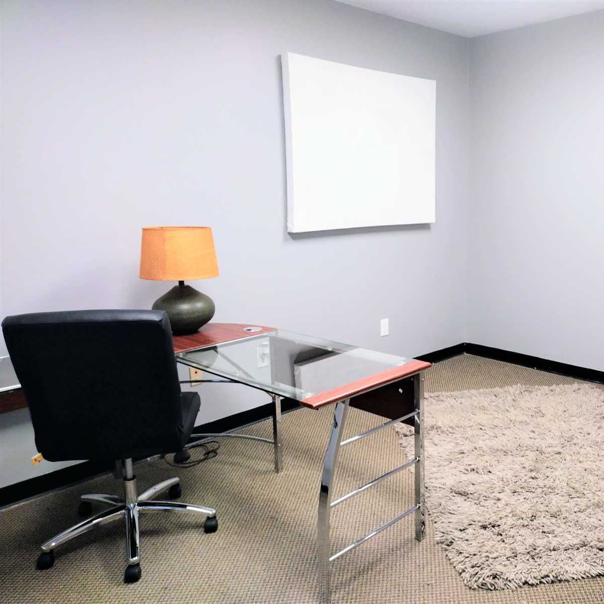 A minimalist office space featuring a glass-top desk with a chair, a lamp on the desk, and a large area rug on the floor.