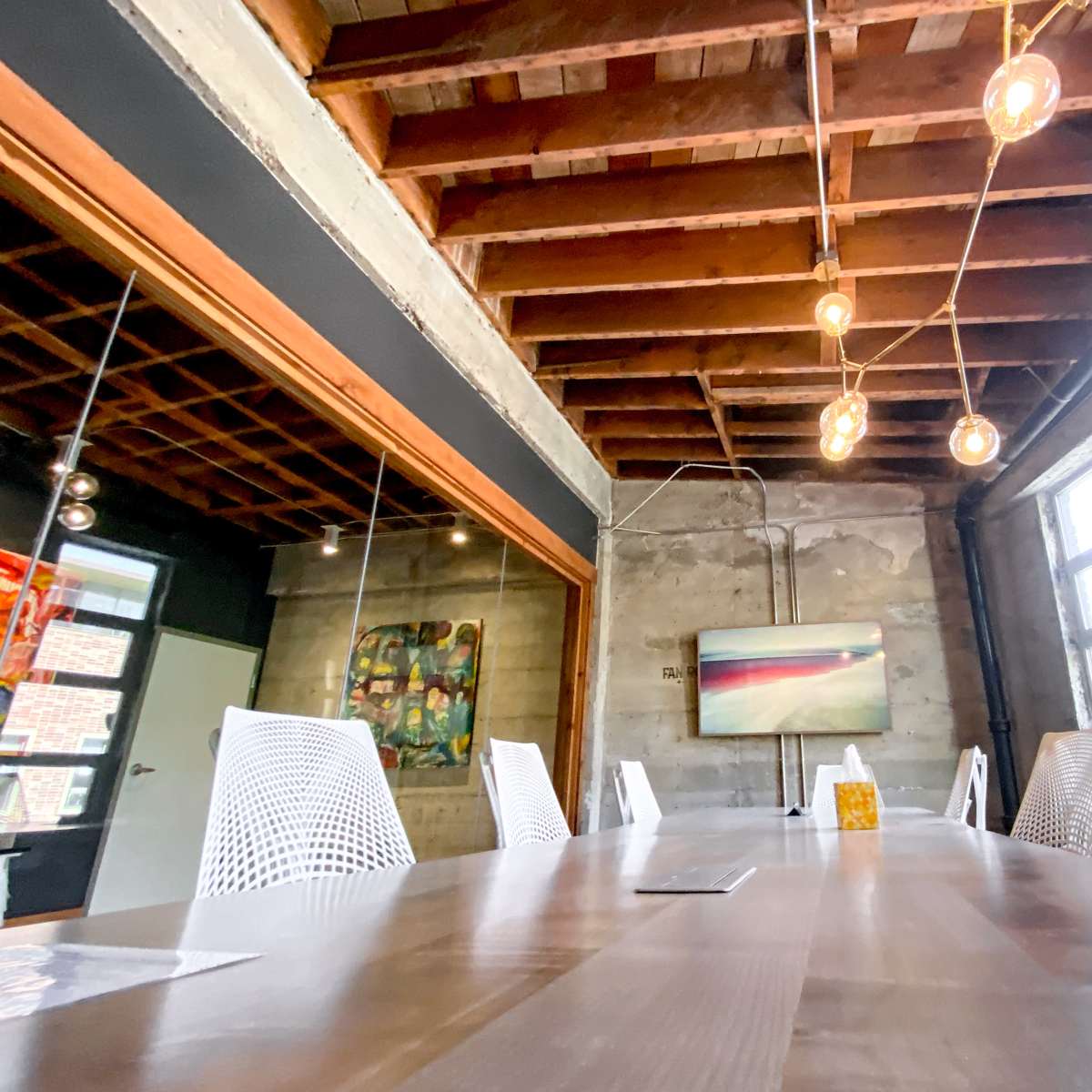 A long wooden table surrounded by white chairs is set in a room with exposed wooden beams and concrete walls, featuring large windows and a mounted television.