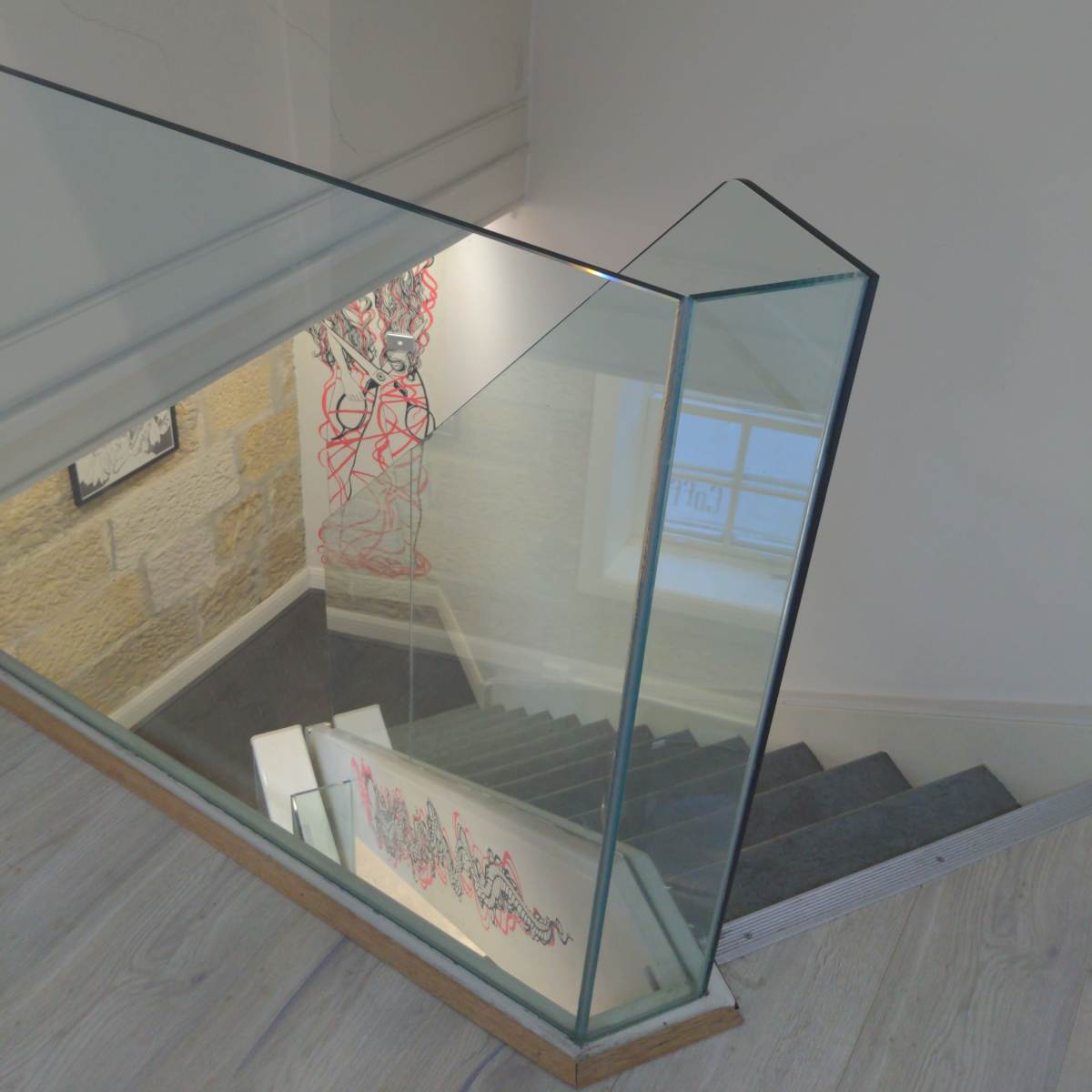 A glass railing overlooking a stairway with light-colored wooden steps, leading down to a room with artwork visible on the walls.