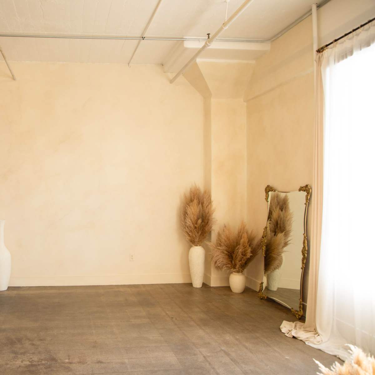 A minimalist room with a pale wall, a fireplace with a mirror above it, and decorative elements like vases and tall dried plants.