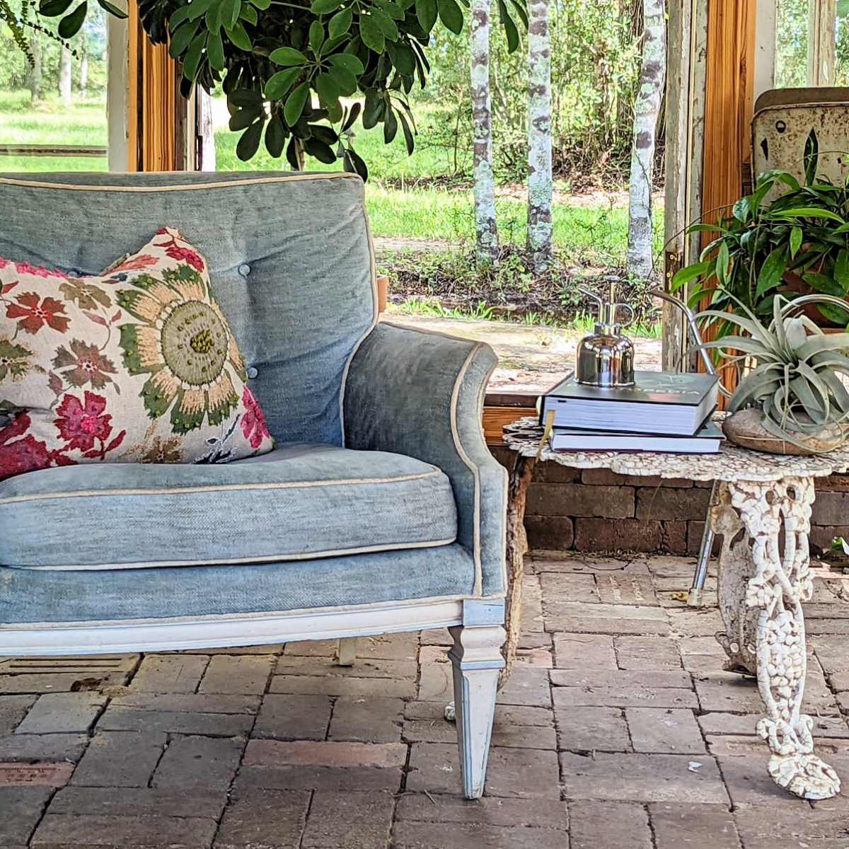 A light blue armchair with a decorative pillow sits beside a small, ornate side table with a potted plant and a silver container in a sunlit interior space.