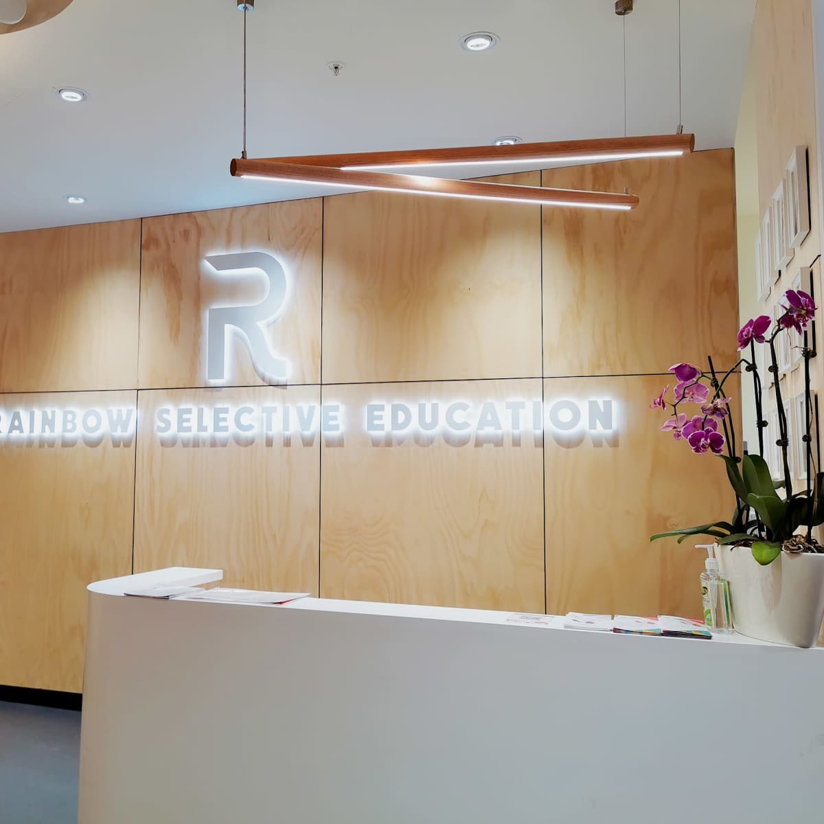 The reception area of an educational center with a wooden wall featuring a glowing "RAINBOW SELECTIVE EDUCATION" sign and a curved front desk.