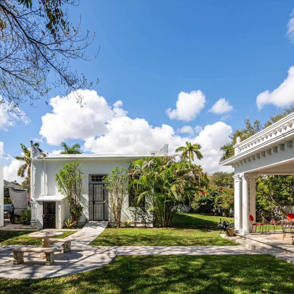 A landscaped outdoor area featuring a white building, a pavilion with columns, and a circular stone bench surrounded by greenery under a partly cloudy sky.