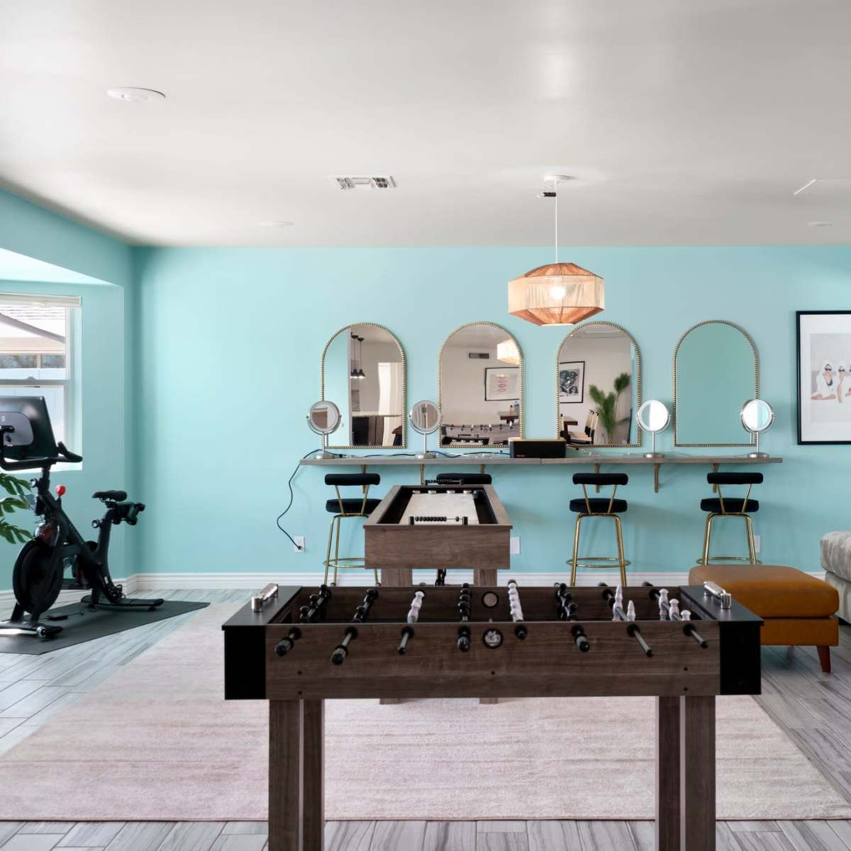 A game room features a foosball table in the foreground, a bar with stools in the background, and a seating area with a couch and decorative elements.
