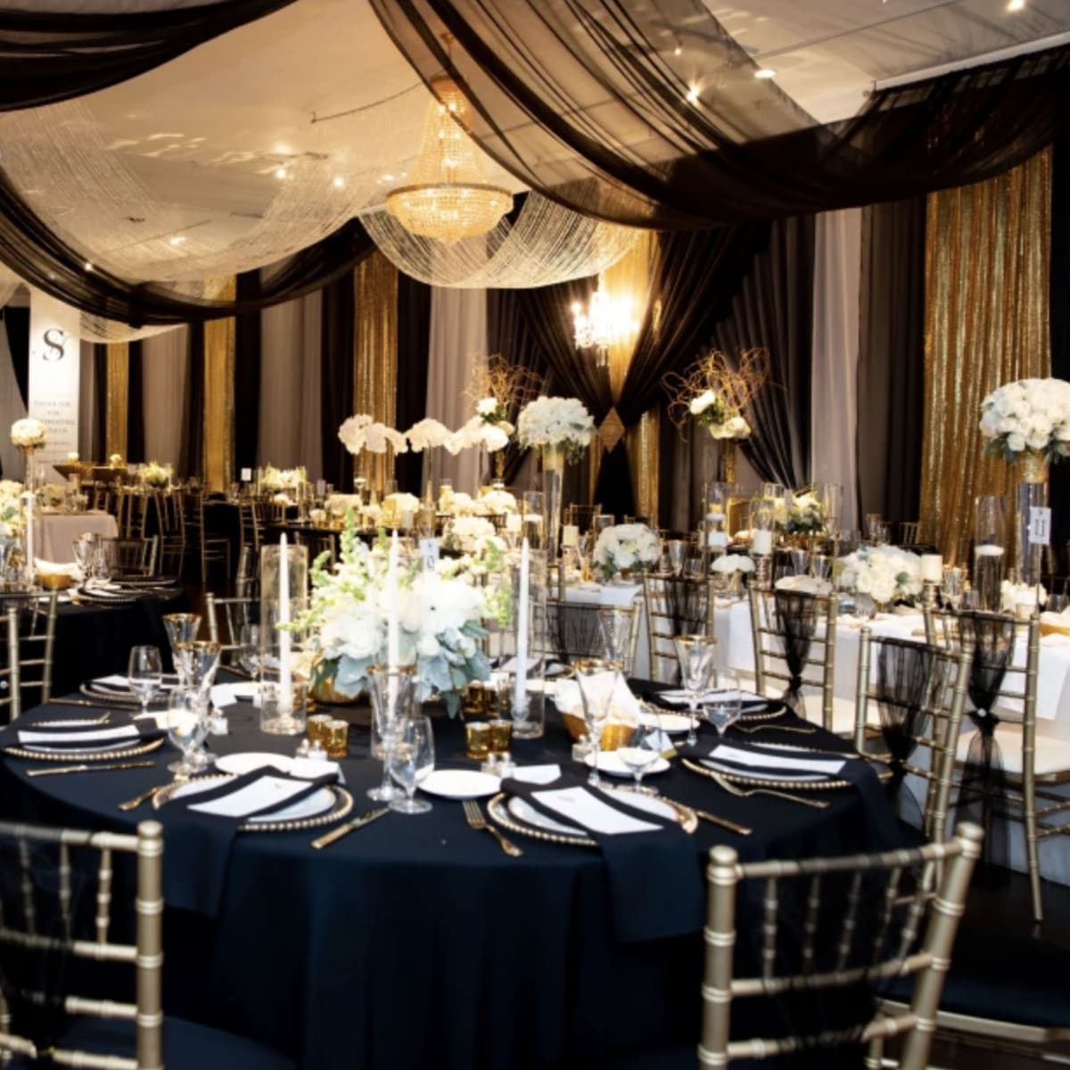 A elegantly decorated banquet hall with round tables set for a formal event, featuring floral centerpieces and draped fabric overhead.