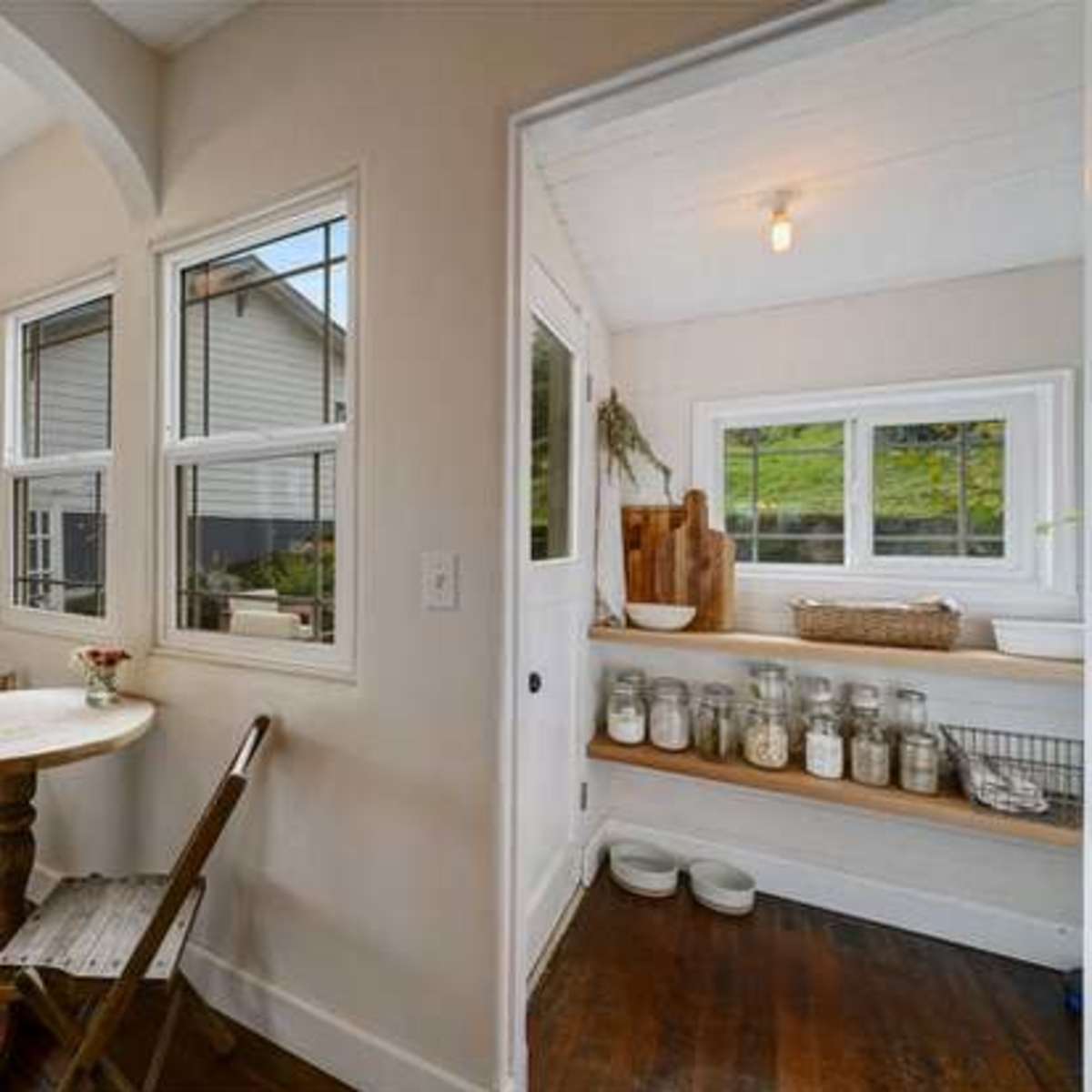 A small kitchen space features a round table with a single chair, a chalkboard on the wall, and a windowed nook displaying jars and a wooden cutting board.