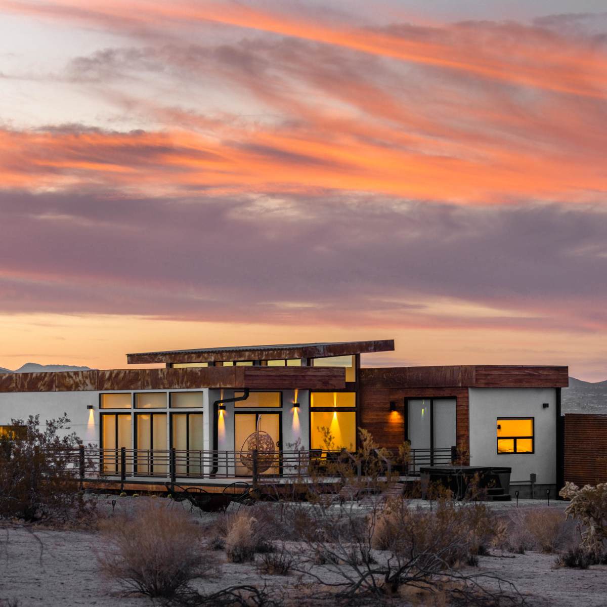 A modern house with large windows and a wooden deck is set against a colorful sunset in a desert landscape.