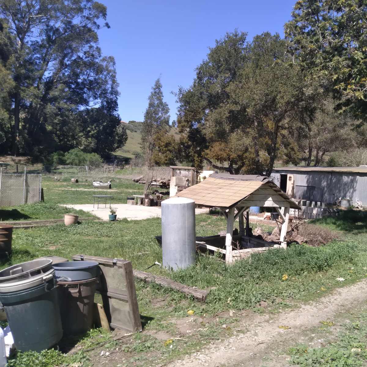 A rural scene with a small wooden structure, a storage tank, and various containers on a grassy area near a mobile home.