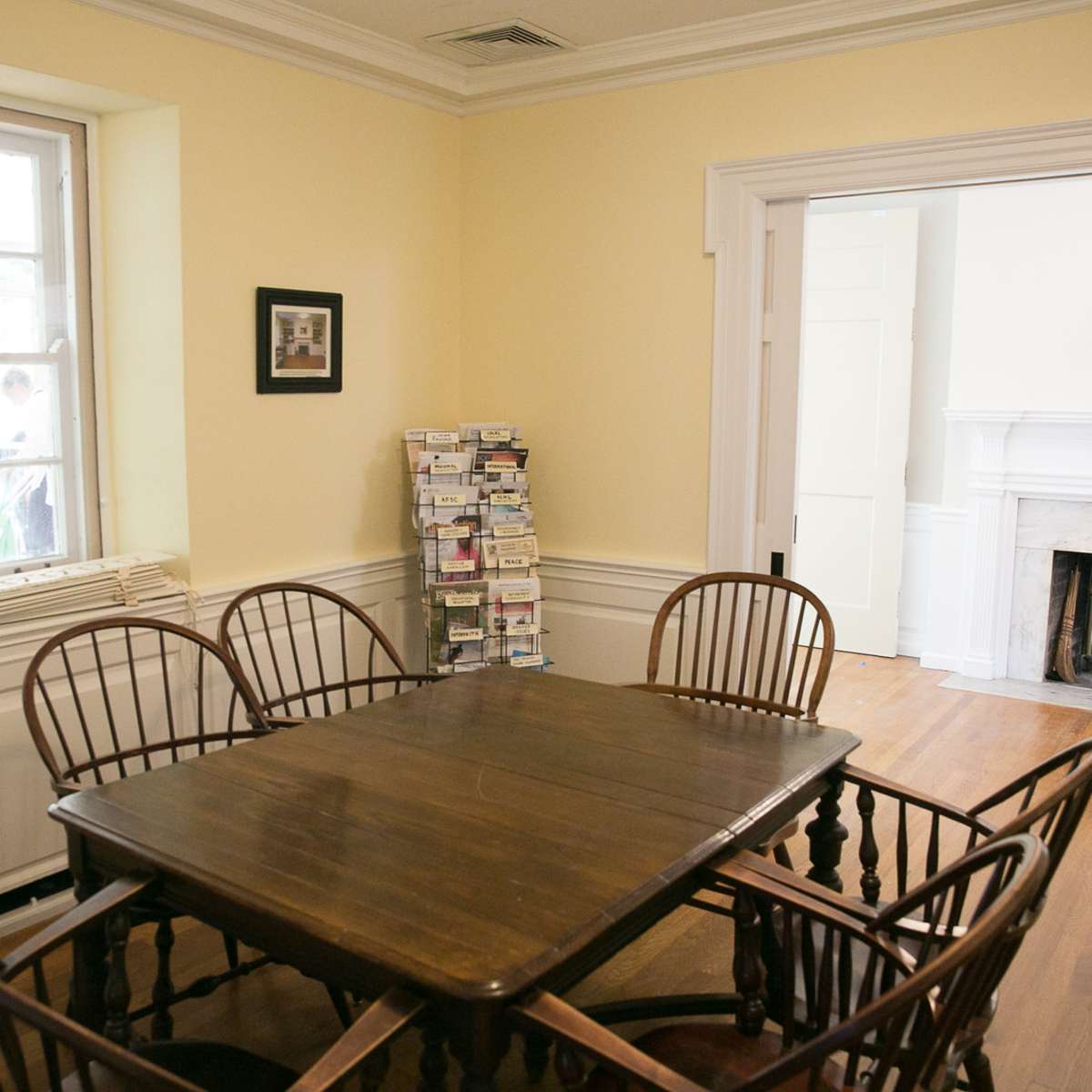 A wooden table surrounded by six chairs is positioned in a light-colored room with a window, a bookshelf, and a fireplace.