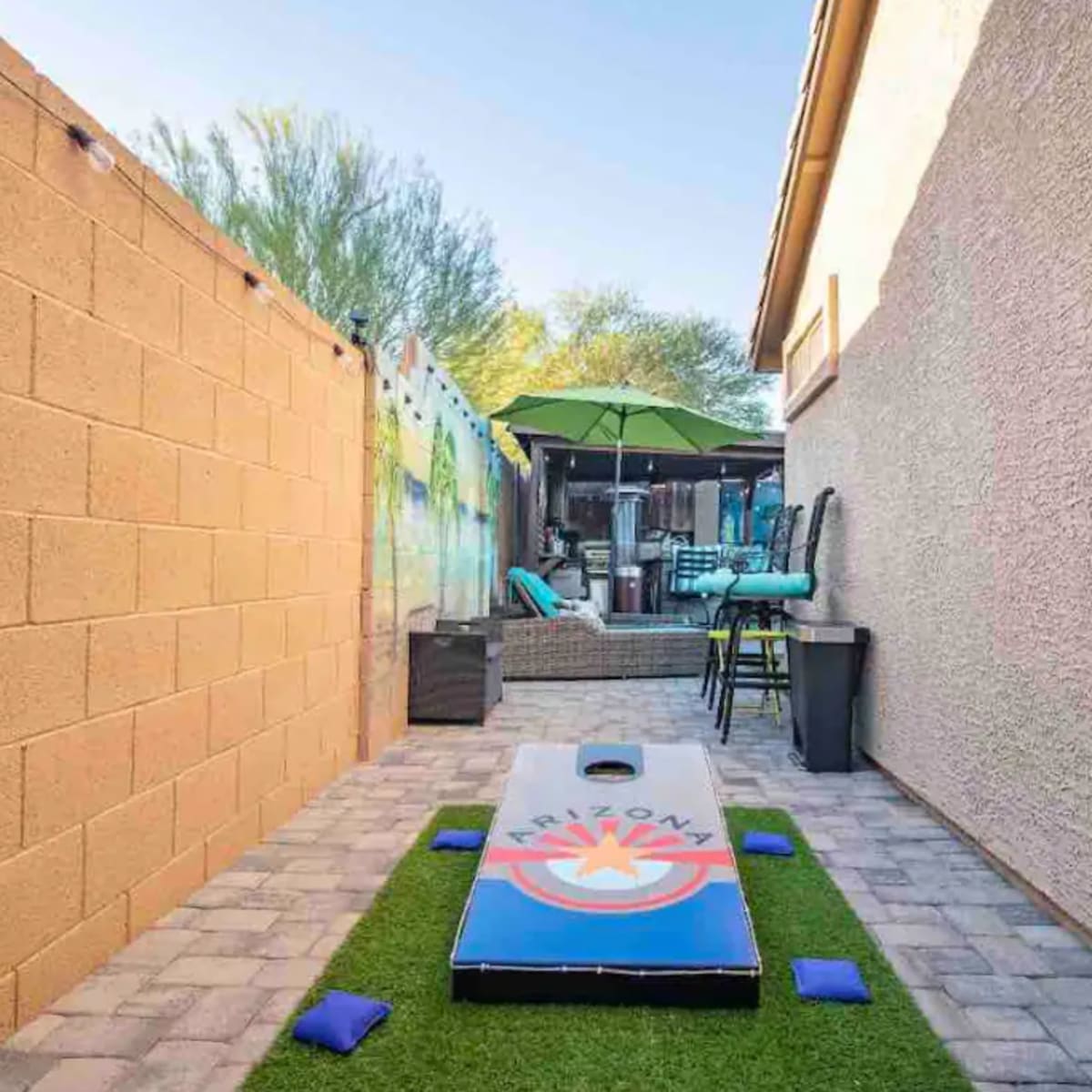 A narrow outdoor space featuring a cornhole game board on a patch of artificial grass, surrounded by walls and patio furniture under a green umbrella.
