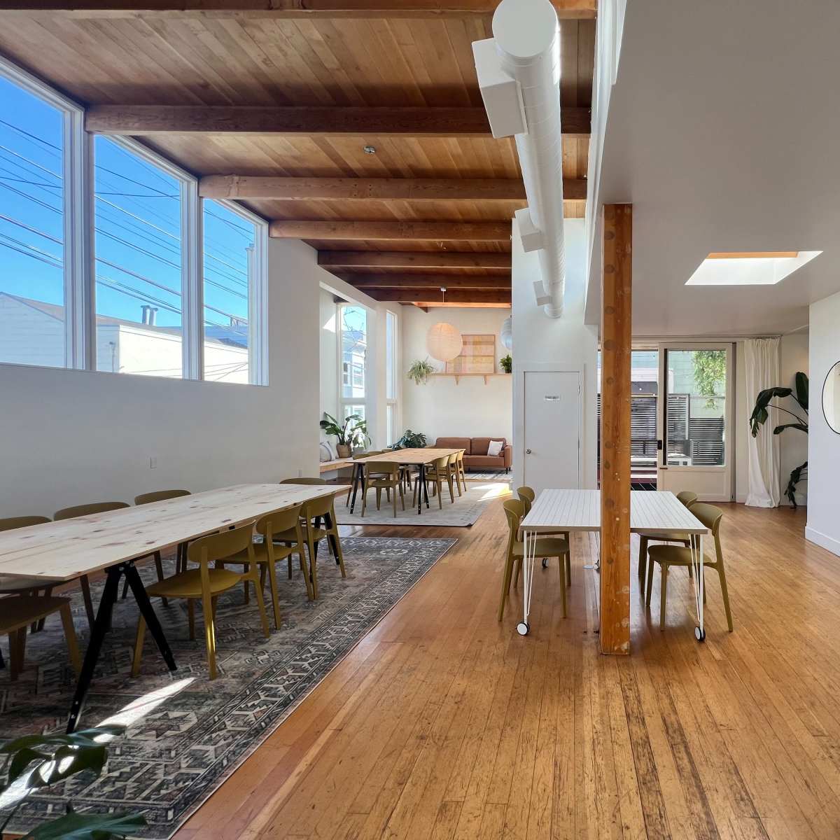 A spacious, modern interior with wooden beams, tables arranged for dining or meetings, and large windows allowing natural light.