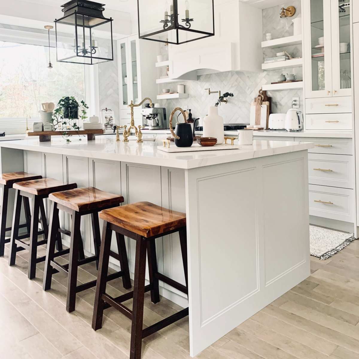 A modern kitchen features a central island with four wooden stools, pendant lights, and white cabinetry against a marble backsplash.