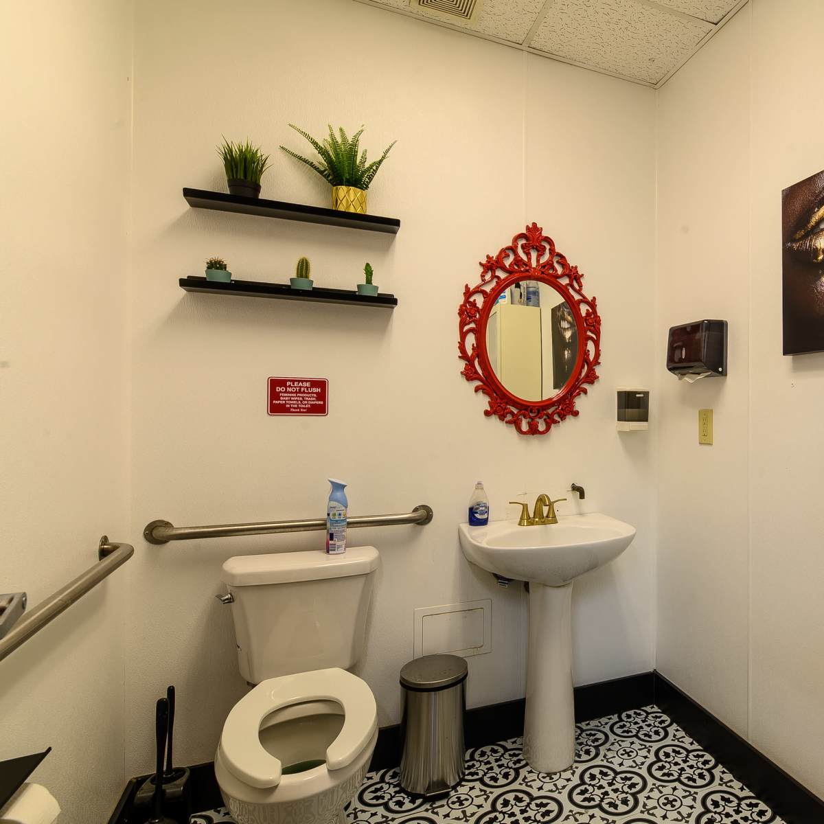 A small bathroom featuring a toilet, a sink with a mirror, a wall-mounted hand sanitizer dispenser, and decorative shelves with plants.