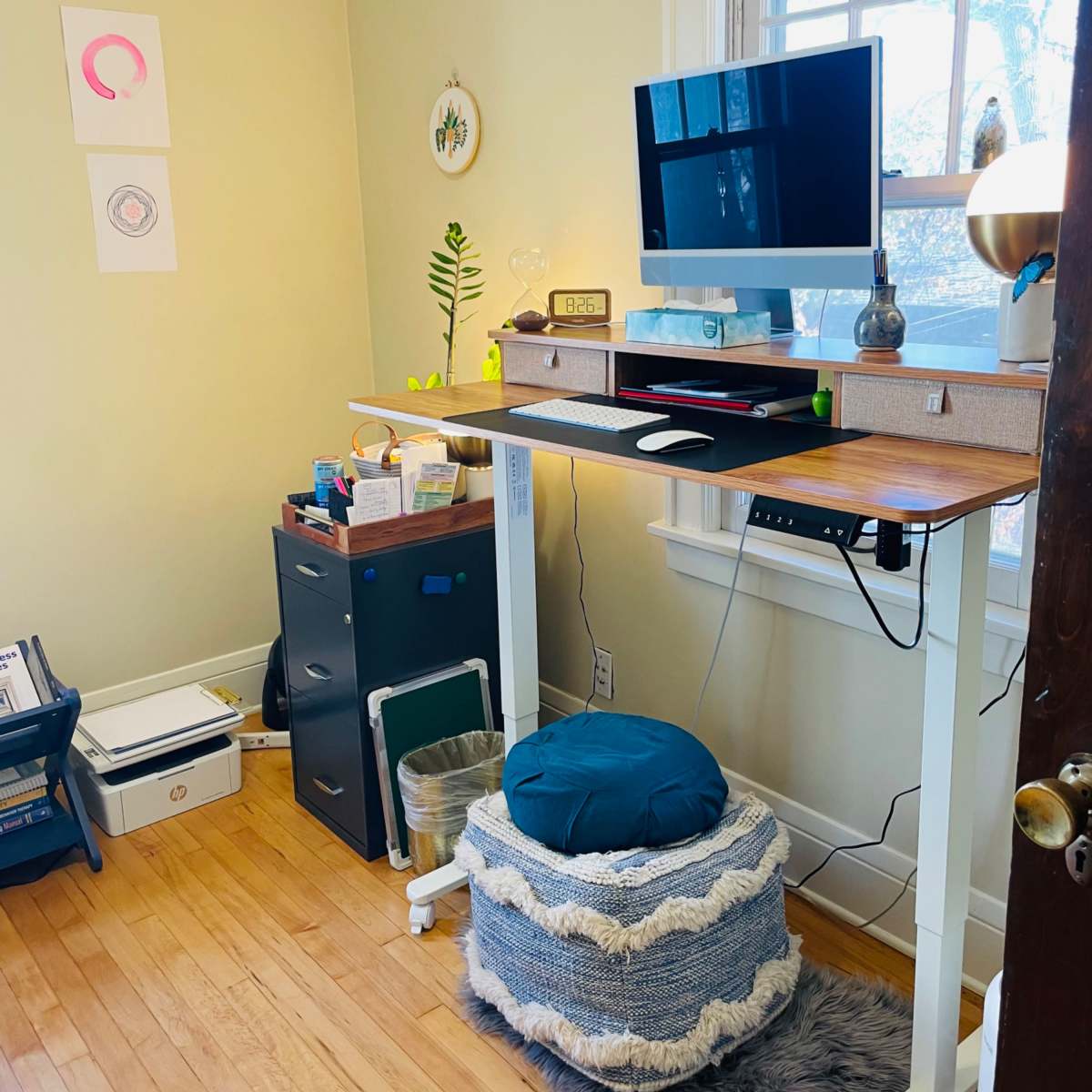 A standing desk with a monitor and lamp is positioned against a window in a room featuring a storage cabinet and decorative items on the walls.