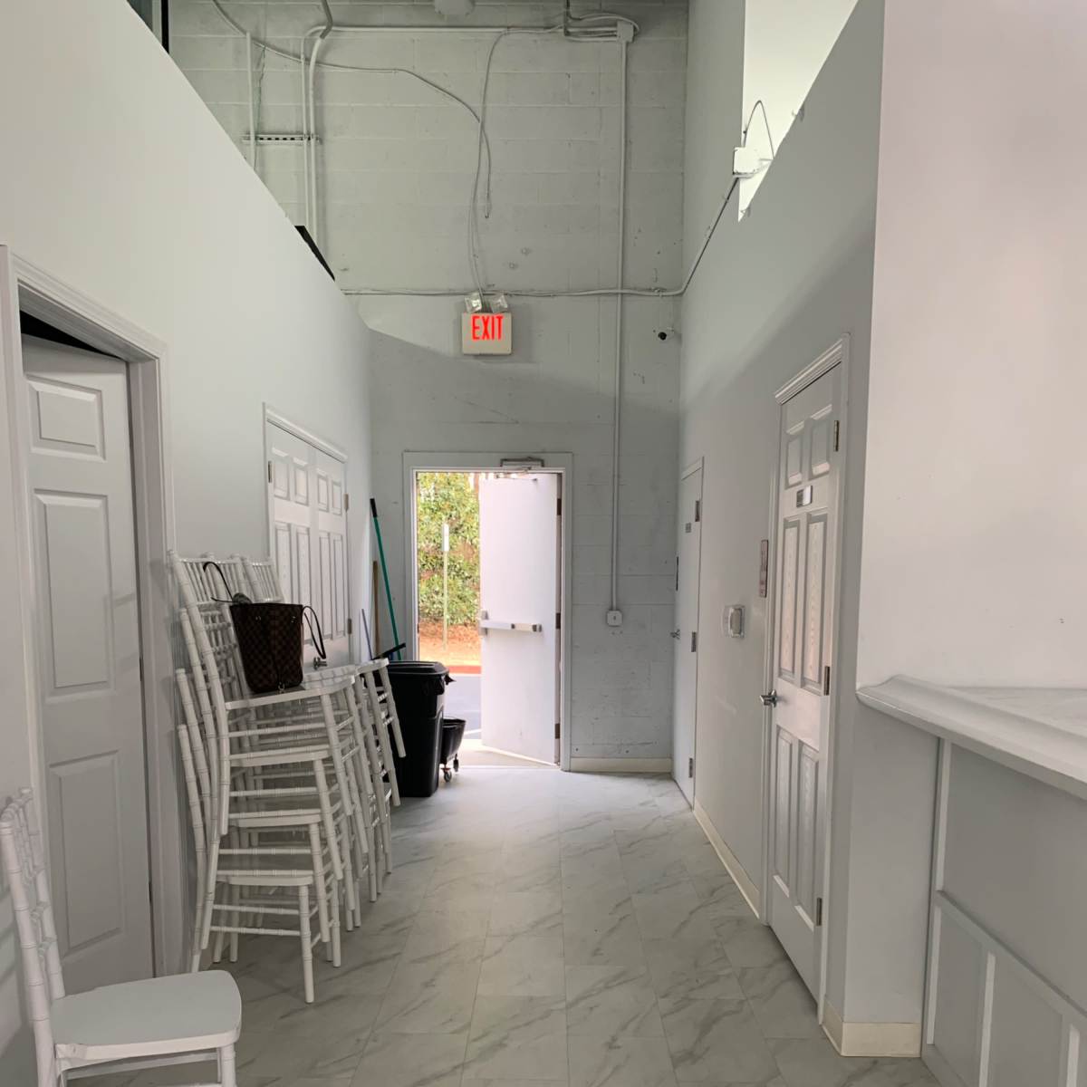 A long hallway with white walls, chairs stacked against one side, and a door leading outside at the end.
