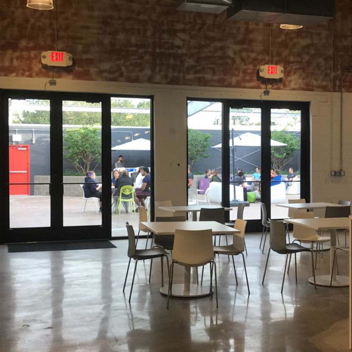 A modern indoor space with tables and chairs, overlooking an outdoor area where people are sitting under umbrellas.